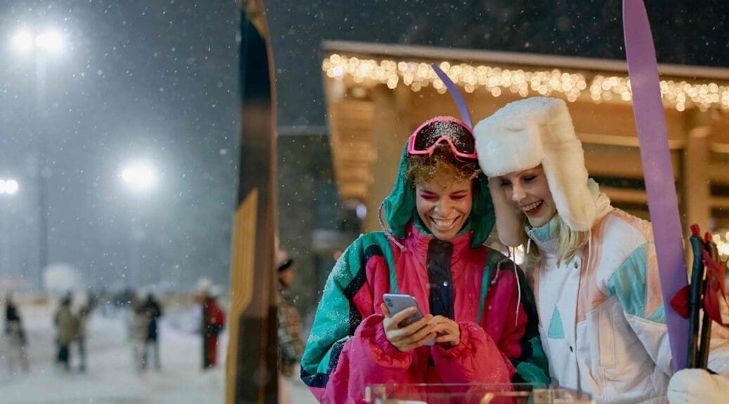 Two female students wearing ski gear laugh at a phone in the Alps